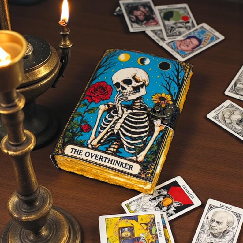 May include: A decorative journal with a skeleton illustration on the cover, the text "THE OVERTHINKER" is displayed below the illustration. The journal is surrounded by tarot cards and lit candles, creating a mystical ambiance.