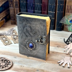 May include: A gray leather journal with a dragon embossed on the cover. The journal has a metal clasp and a blue gemstone set in the center of the dragon's chest.
