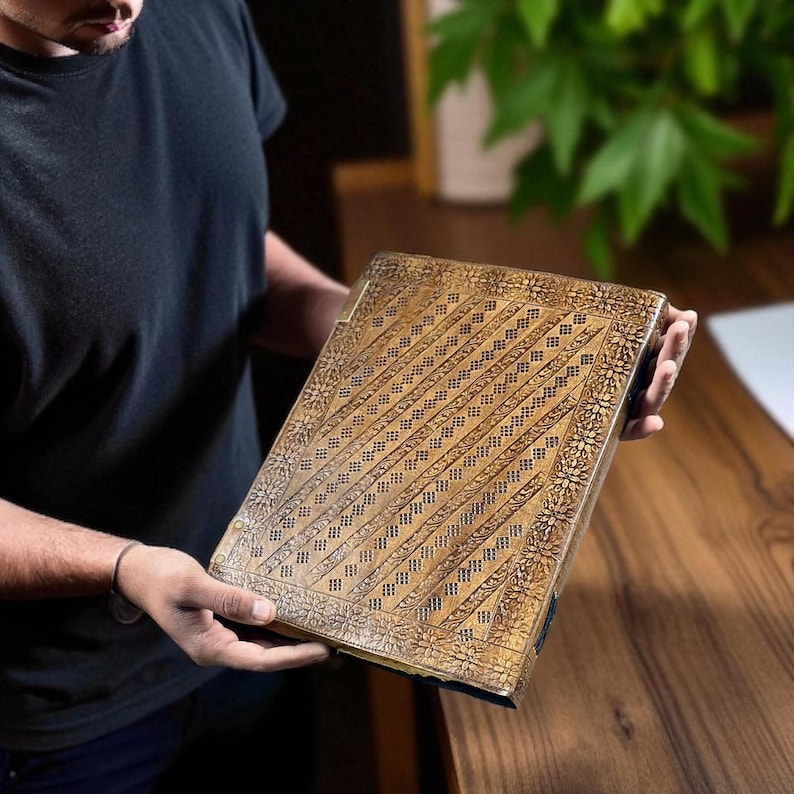 Pode incluir: Um livro retangular em estilo antigo com um design entalhado detalhado. A capa &eacute; marrom dourado quente com padr&otilde;es diagonais e bordas florais. O livro &eacute; segurado por uma pessoa, mostrando seu tamanho e artesanato.