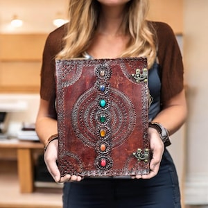 Puede incluir: Un diario de cuero marr&oacute;n hecho a mano con un dise&ntilde;o en relieve decorativo y una fila de piedras preciosas de colores. El diario tiene protectores de esquina de metal y un cierre de broche. La cubierta est&aacute; adornada con patrones intrincados.