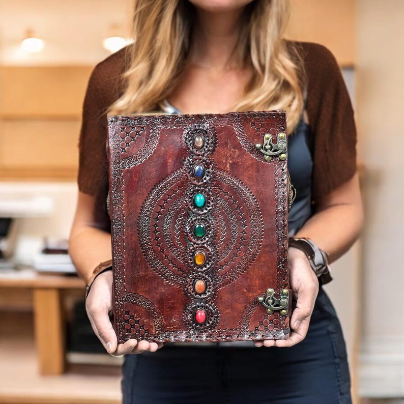 Puede incluir: Un diario de cuero marr&oacute;n hecho a mano con un dise&ntilde;o en relieve decorativo y una fila de piedras preciosas de colores. El diario tiene protectores de esquina de metal y un cierre de broche. La cubierta est&aacute; adornada con patrones intrincados.