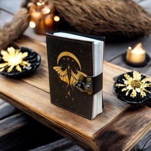 Puede incluir: Un diario de cuero negro con un diseño dorado de polilla y luna creciente. El diario tiene un cierre de broche y una correa de cuero negro. Las páginas son blancas. El diario está sobre una bandeja de madera con elementos decorativos.