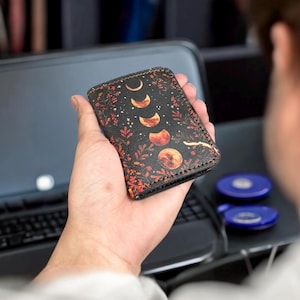 K&ouml;nnte beinhalten: Ein schwarzer Kartenhalter aus Leder mit einer vertikalen Abfolge von Mondphasen in Orange- und Gelbt&ouml;nen, umgeben von einem detaillierten botanischen Druck aus roten und orangefarbenen Bl&auml;ttern und kleinen Sternen, mit sichtbaren dunklen N&auml;hten.