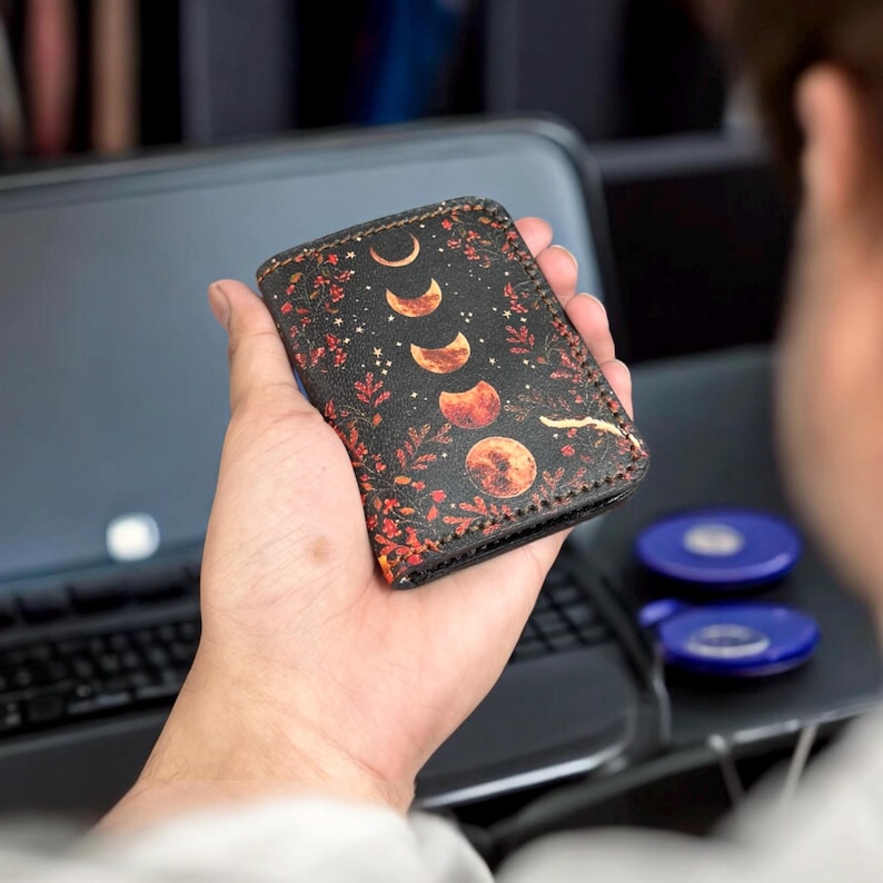 K&ouml;nnte beinhalten: Ein schwarzer Kartenhalter aus Leder mit einer vertikalen Abfolge von Mondphasen in Orange- und Gelbt&ouml;nen, umgeben von einem detaillierten botanischen Druck aus roten und orangefarbenen Bl&auml;ttern und kleinen Sternen, mit sichtbaren dunklen N&auml;hten.