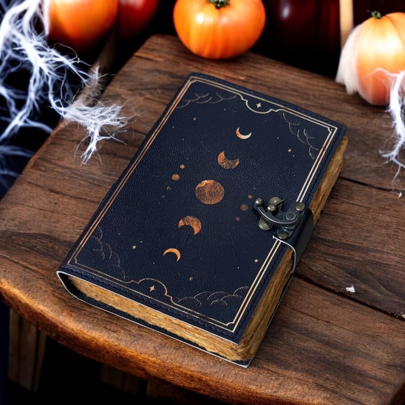 K&ouml;nnte beinhalten: Ein schwarzes Lederjournal mit einem goldenen Mond- und Sternenmuster. Das Journal hat einen Verschluss mit einer Schlie&szlig;e und einen abgenutzten, Vintage-Look.