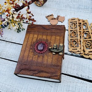 May include: A brown leather journal with a decorative metal clasp and a purple gemstone embedded in the front cover. The journal has a distressed look and is decorated with intricate stitching.