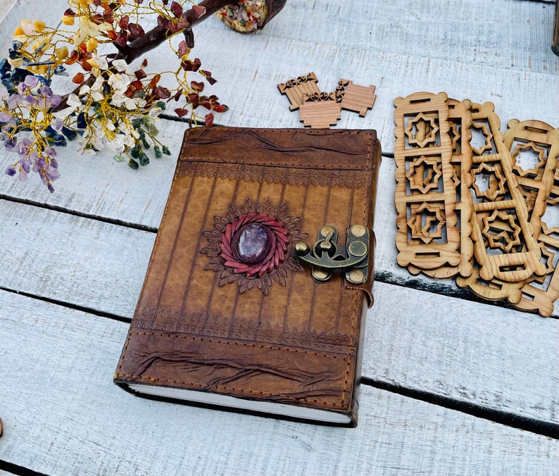 May include: A brown leather journal with a decorative metal clasp and a purple gemstone embedded in the front cover. The journal has a distressed look and is decorated with intricate stitching.