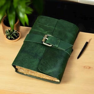 May include: A dark green leather journal with a buckle closure. The journal is closed with a strap and a silver-colored buckle. A black pencil rests on the wooden surface next to the journal. A small potted plant is in the background.