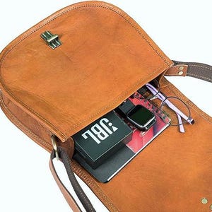 Pu&ograve; includere: Una borsa a tracolla in pelle marrone con chiusura a lembo e fibbia. La borsa &egrave; aperta e contiene un altoparlante JBL nero, un quaderno e un paio di occhiali.