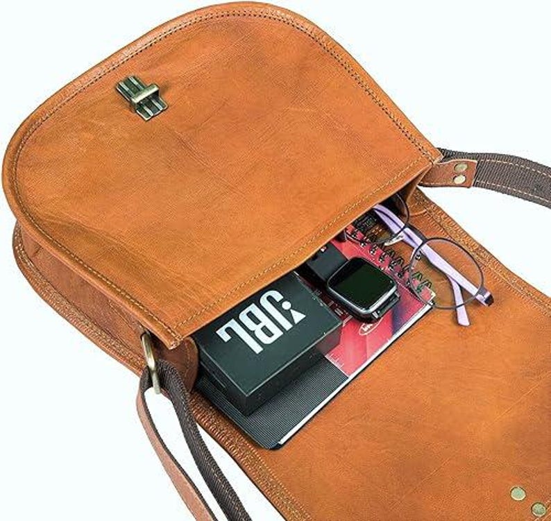 Pu&ograve; includere: Una borsa a tracolla in pelle marrone con chiusura a lembo e fibbia. La borsa &egrave; aperta e contiene un altoparlante JBL nero, un quaderno e un paio di occhiali.