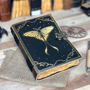 Puede incluir: Un diario de cuero negro con un diseño de polilla dorada y un cierre de metal. El diario tiene un aspecto desgastado y está decorado con estrellas doradas y nubes.