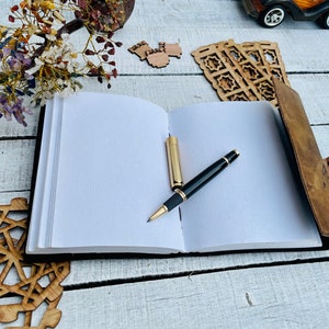 May include: A brown leather-bound journal with a pen resting on the open pages. The journal is on a white wooden surface with decorative wooden pieces around it.