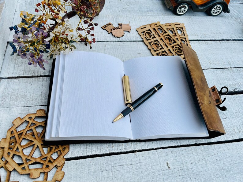 May include: A brown leather-bound journal with a pen resting on the open pages. The journal is on a white wooden surface with decorative wooden pieces around it.