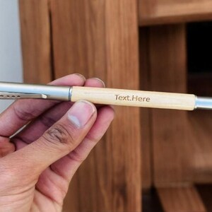 May include: A light-coloured wooden pen with a silver metal clip and tip. The pen has the text "Text.Here" printed on the barrel. The pen is held in a hand against a blurred wooden background.