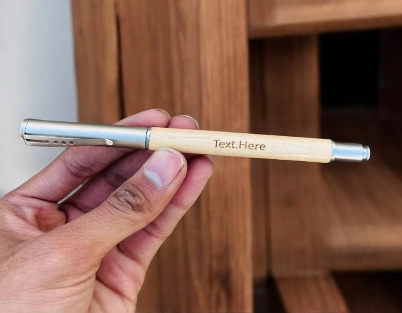May include: A light-coloured wooden pen with a silver metal clip and tip. The pen has the text "Text.Here" printed on the barrel. The pen is held in a hand against a blurred wooden background.