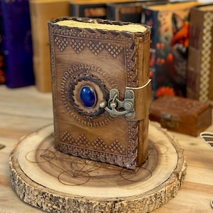 May include: A brown leather journal with a blue gemstone and a brass clasp. The journal has intricate embossed designs and is sitting on a wooden surface.