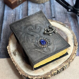 May include: A black leather journal with a dragon design embossed on the cover. The journal has a metal clasp with a blue gemstone. The journal is sitting on a wooden surface.