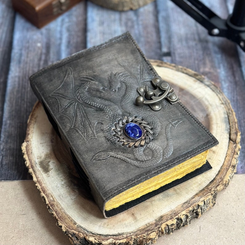 May include: A black leather journal with a dragon design embossed on the cover. The journal has a metal clasp with a blue gemstone. The journal is sitting on a wooden surface.