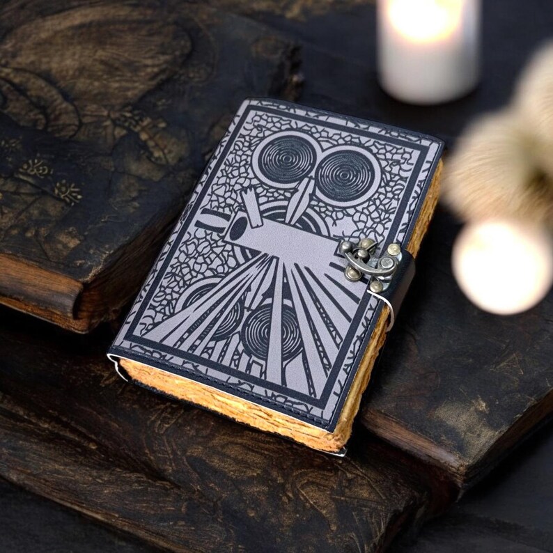 May include: A leather-bound journal with a silver geometric design on the cover. The journal has a metal clasp and aged, yellowed pages. The cover design features circular and linear patterns. The journal is surrounded by other antique books.