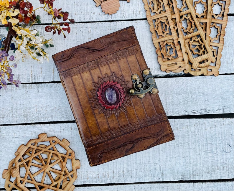 May include: A brown leather journal with a decorative floral design and a purple gemstone in the center. The journal has a metal clasp and is closed.