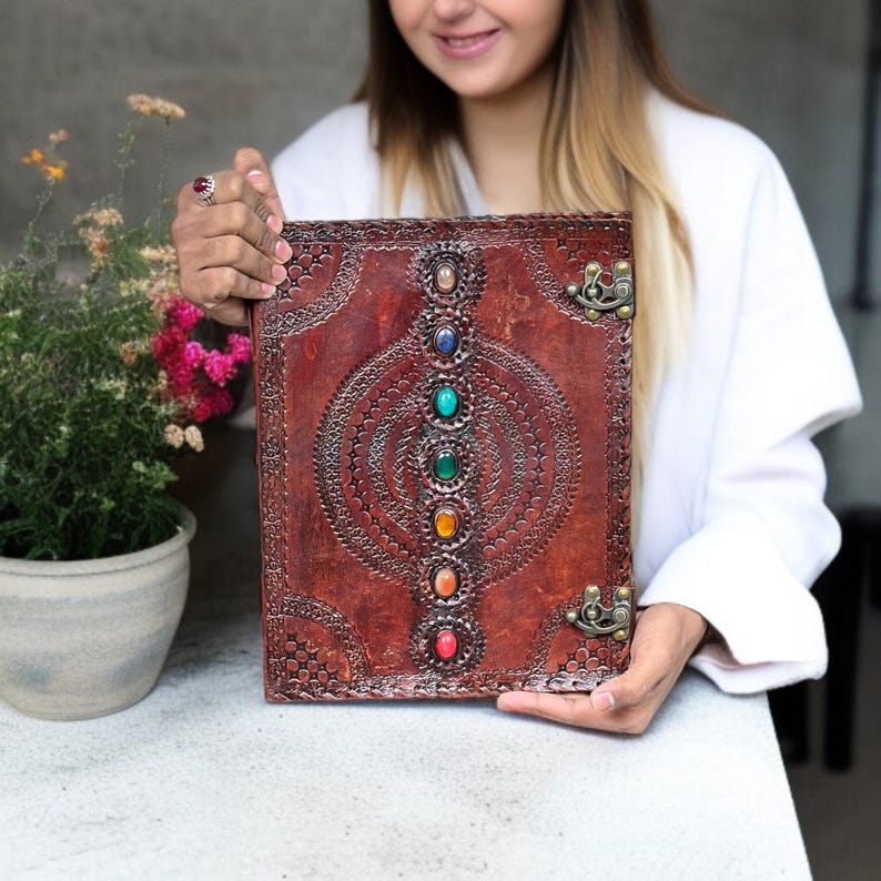 Puede incluir: Un diario de cuero marr&oacute;n con un dise&ntilde;o en relieve decorativo y una fila de piedras preciosas de colores. El diario tiene un cierre de broche, mostrando los intrincados detalles.