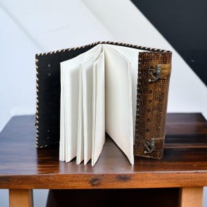 Puede incluir: Un diario abierto de estilo vintage con p&aacute;ginas en blanco. El diario presenta una cubierta marr&oacute;n oscuro en relieve con un cierre y una lomo de cuero negro con costuras color crema. Las p&aacute;ginas son de color crema.