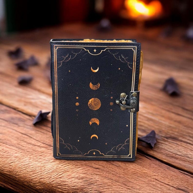 K&ouml;nnte beinhalten: Ein schwarzes Lederjournal mit einem goldenen Mondphasen-Design und einem Messingverschluss. Das Journal hat einen Used-Look und ist mit goldenen Sternen verziert.