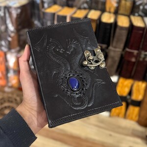 May include: Black leather journal with a detailed embossed design of two dragons intertwined. The journal has a blue gemstone set in the center of the design and a brass clasp.