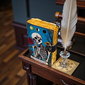 May include: A leather-bound journal with a blue cover featuring a skeleton illustration, a red rose, and the text "THE OVERTHINKER." The journal has aged, yellowed pages and a black strap closure. A white feather quill sits nearby.