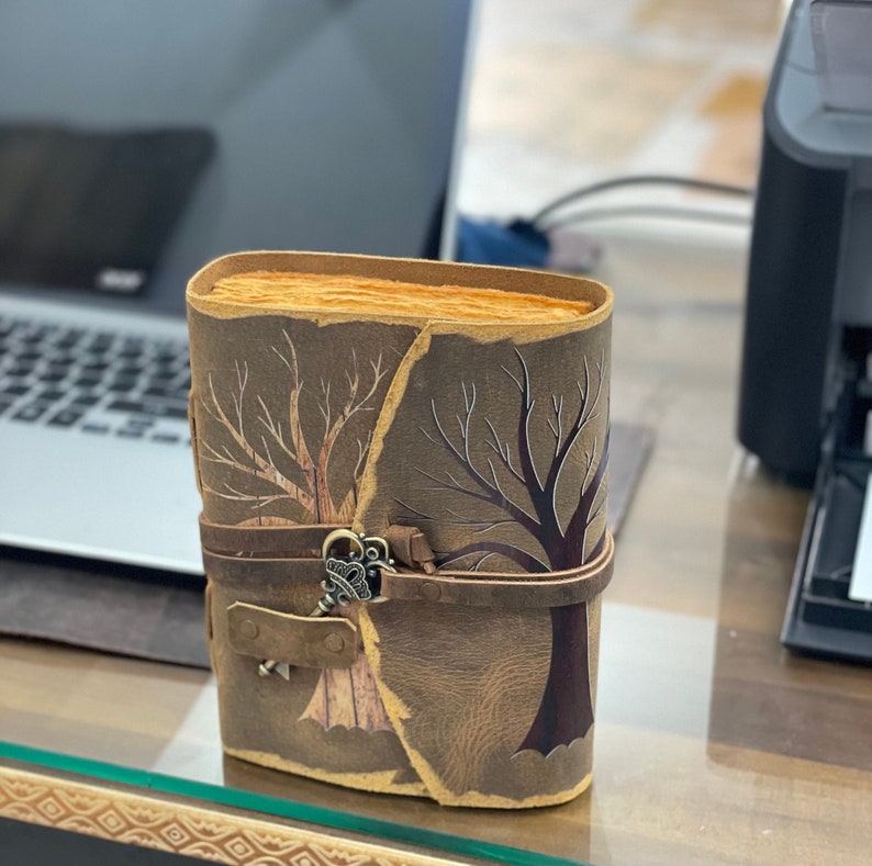 May include: A brown leather journal with a keyhole design and a metal key charm. The journal has two trees etched into the front cover, one on each side of the keyhole.