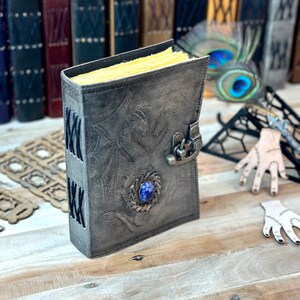 May include: A gray leather journal with a dragon design and a blue gemstone. The journal has a clasp closure and is closed.