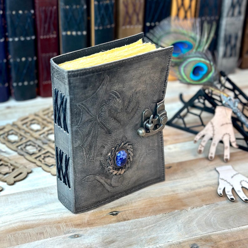 May include: A gray leather journal with a dragon design and a blue gemstone. The journal has a clasp closure and is closed.