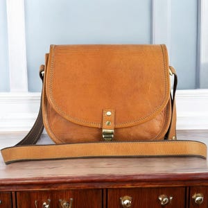 Pu&ograve; includere: Una borsa a tracolla in pelle marrone con chiusura a lembo e una lunga tracolla regolabile. La borsa ha un aspetto usurato e vintage.