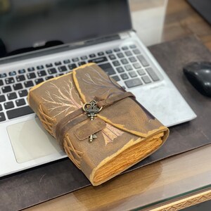 May include: A brown leather journal with a tree design and a decorative key charm. The journal is closed and sitting on a laptop.