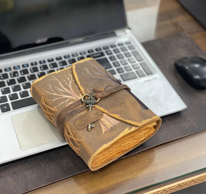 May include: A brown leather journal with a tree design and a decorative key charm. The journal is closed and sitting on a laptop.