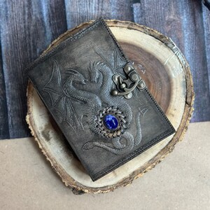 May include: A dark gray leather journal with a tooled design of two intertwined dragons and a blue gemstone clasp.