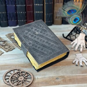 May include: A gray leather-bound journal with a decorative embossed pattern. The journal has yellowed pages and is closed. The journal is sitting on a wooden surface.