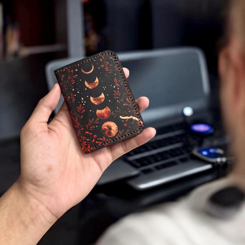 K&ouml;nnte beinhalten: Ein rechteckiges Kartenetui aus schwarzem Leder mit einem Aufdruck der Mondphasen in Orange- und Goldt&ouml;nen, umgeben von kleinen Sternen und roten Pflanzenmotiven, mit sichtbaren dunklen N&auml;hten entlang der Kanten des Etuis.