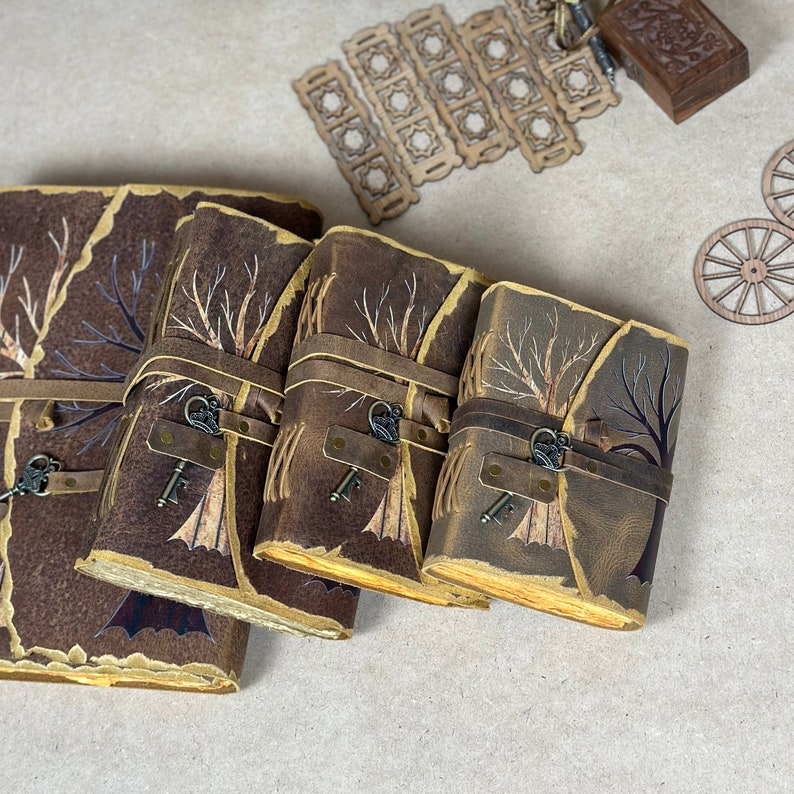 May include: Four brown leather journals with a tree design and a key charm. The journals are stacked on top of each other, with the largest journal on the bottom and the smallest journal on top.