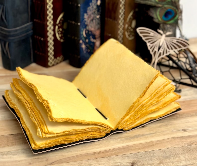 May include: A handmade journal with a black leather cover and yellowed paper pages. The journal is open to reveal the layered pages.