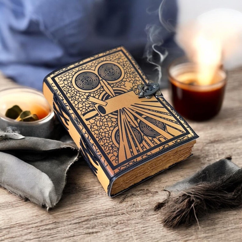 May include: A tan leather journal with a detailed black art deco design on the cover. The journal has a metal clasp and aged, yellowed pages. A small amount of smoke rises from the journal, suggesting it is warm.