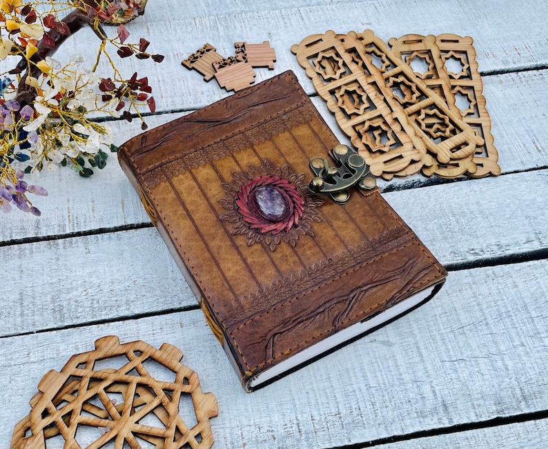 May include: A brown leather journal with a decorative floral design and a gemstone accent. The journal has a metal clasp and is closed.