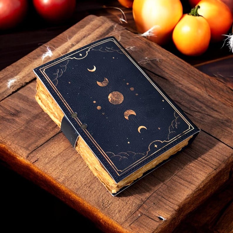 K&ouml;nnte beinhalten: Ein schwarzes, ledergebundenes Tagebuch mit goldenen Akzenten und einem Mond- und Sternenmuster. Das Tagebuch hat einen abgenutzten, Vintage-Look.