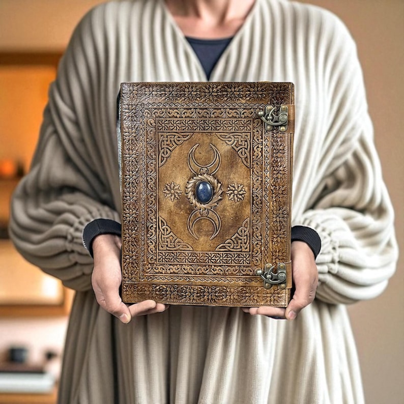 Pode incluir: Um di&aacute;rio de couro castanho com intrincados desenhos em relevo e uma pedra preciosa azul no centro. O di&aacute;rio apresenta um fecho de metal e um padr&atilde;o detalhado nas bordas. A capa inclui motivos de lua crescente e estrelas.