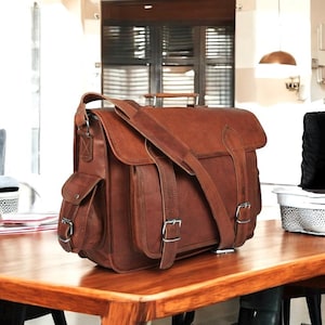 Może przedstawiać: Brązowa sk&oacute;rzana torba listonoszka z dużą kieszenią z przodu i dwoma regulowanymi paskami. Torba ma g&oacute;rny uchwyt i długi pasek na ramię.