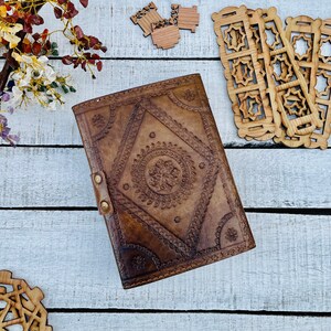 May include: A brown leather journal with intricate embossed designs. The cover features a central diamond-shaped pattern with a circular design inside. The journal is closed and lying on a white wooden surface.
