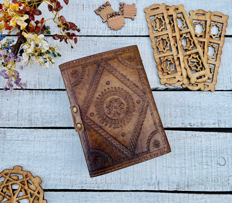 May include: A brown leather journal with intricate embossed designs. The cover features a central diamond-shaped pattern with a circular design inside. The journal is closed and lying on a white wooden surface.