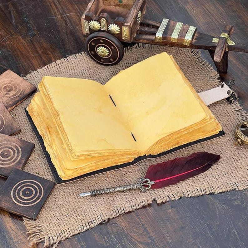 May include: An open, antique-style journal with aged, yellowed pages. A decorative feather quill pen with a burgundy feather rests beside the journal. Wooden coasters and a small wooden cart are also visible.
