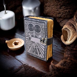 May include: A vintage journal featuring an owl design in black and silver. The pages are aged and yellowed, with a black leather strap and metal clasp. The journal rests on a dark wooden surface.