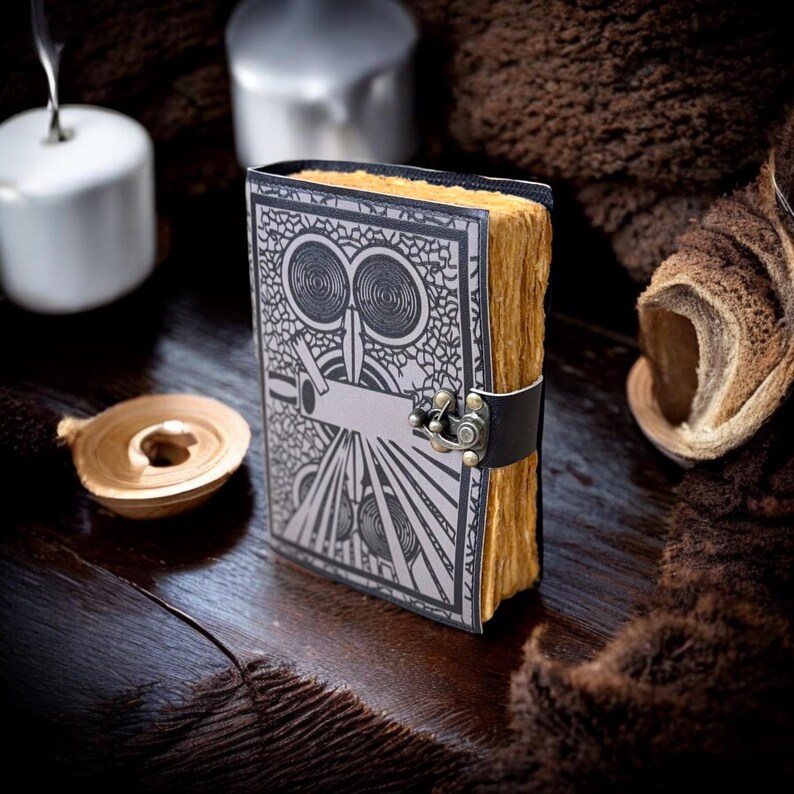 May include: A vintage journal featuring an owl design in black and silver. The pages are aged and yellowed, with a black leather strap and metal clasp. The journal rests on a dark wooden surface.
